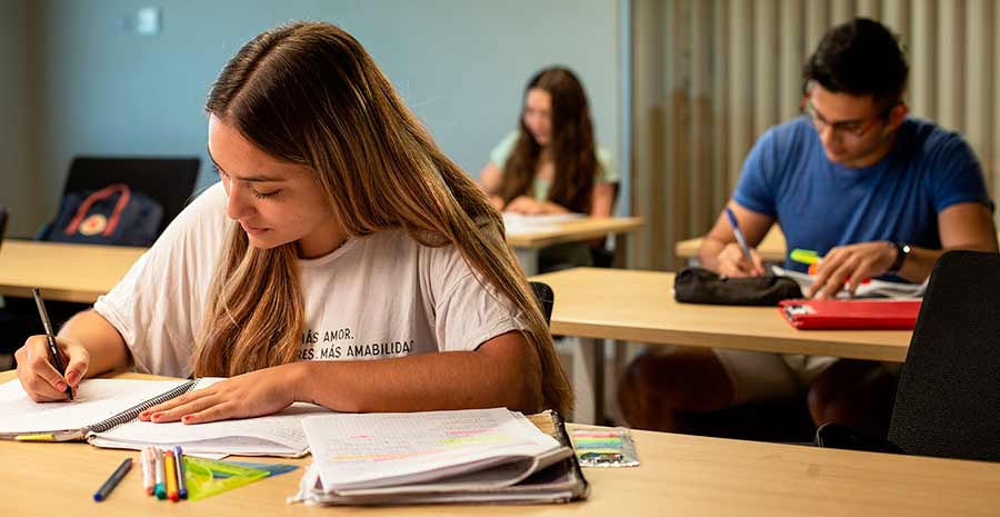 Tres estudiantes en un aula sentados cada uno en su banco, toman apuntes